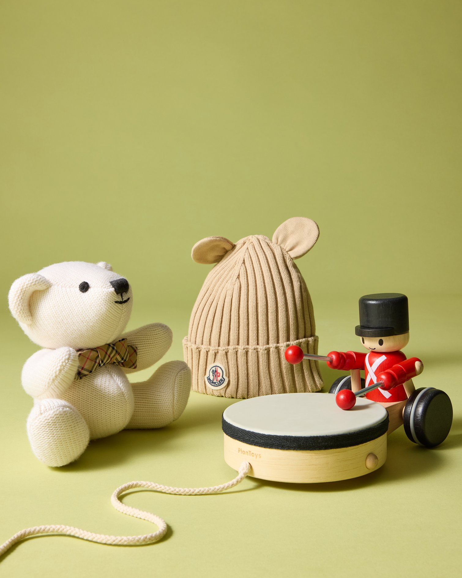 White plush bear, brown ribbed Moncler beanie with ears, and wooden toy soldier with tambourine on a green background