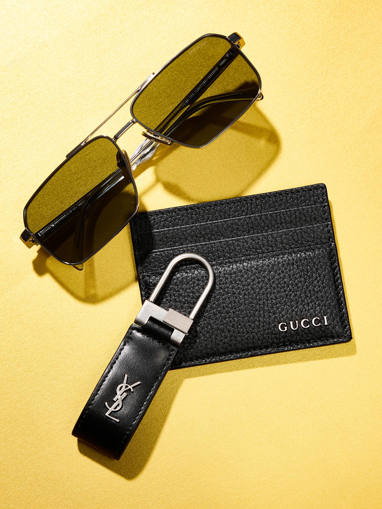 Metal frame sungalsses, black leather Gucci card holder, black leather Saint Laurent key ring against yellow background