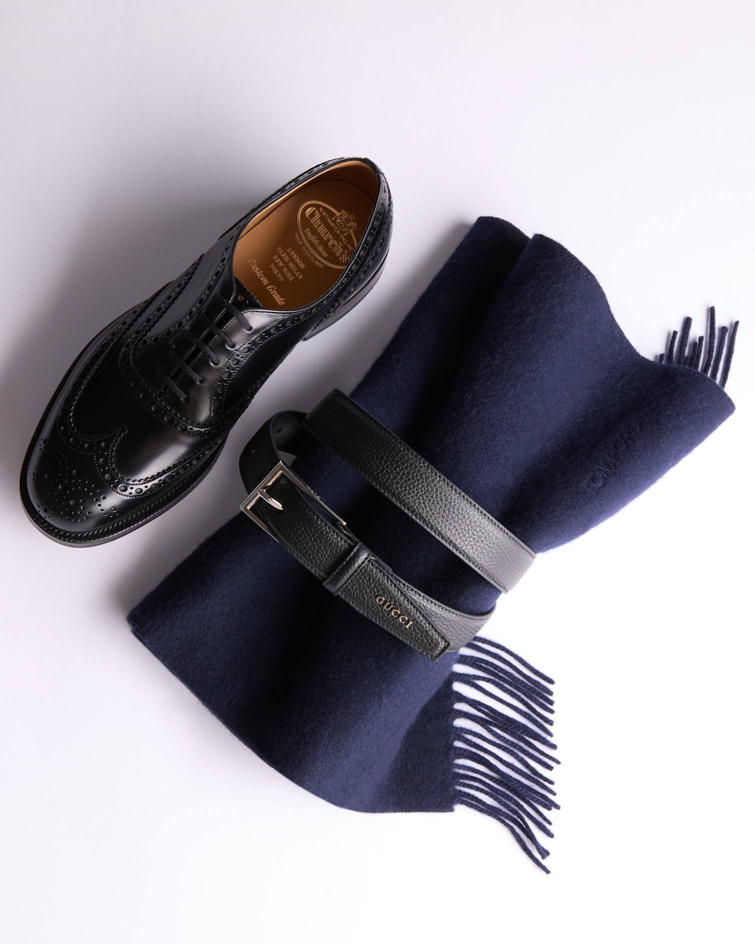 Church’s Black, derby shoe next to Tom Ford scarf with fringe wrapped in a black leather Gucci belt
