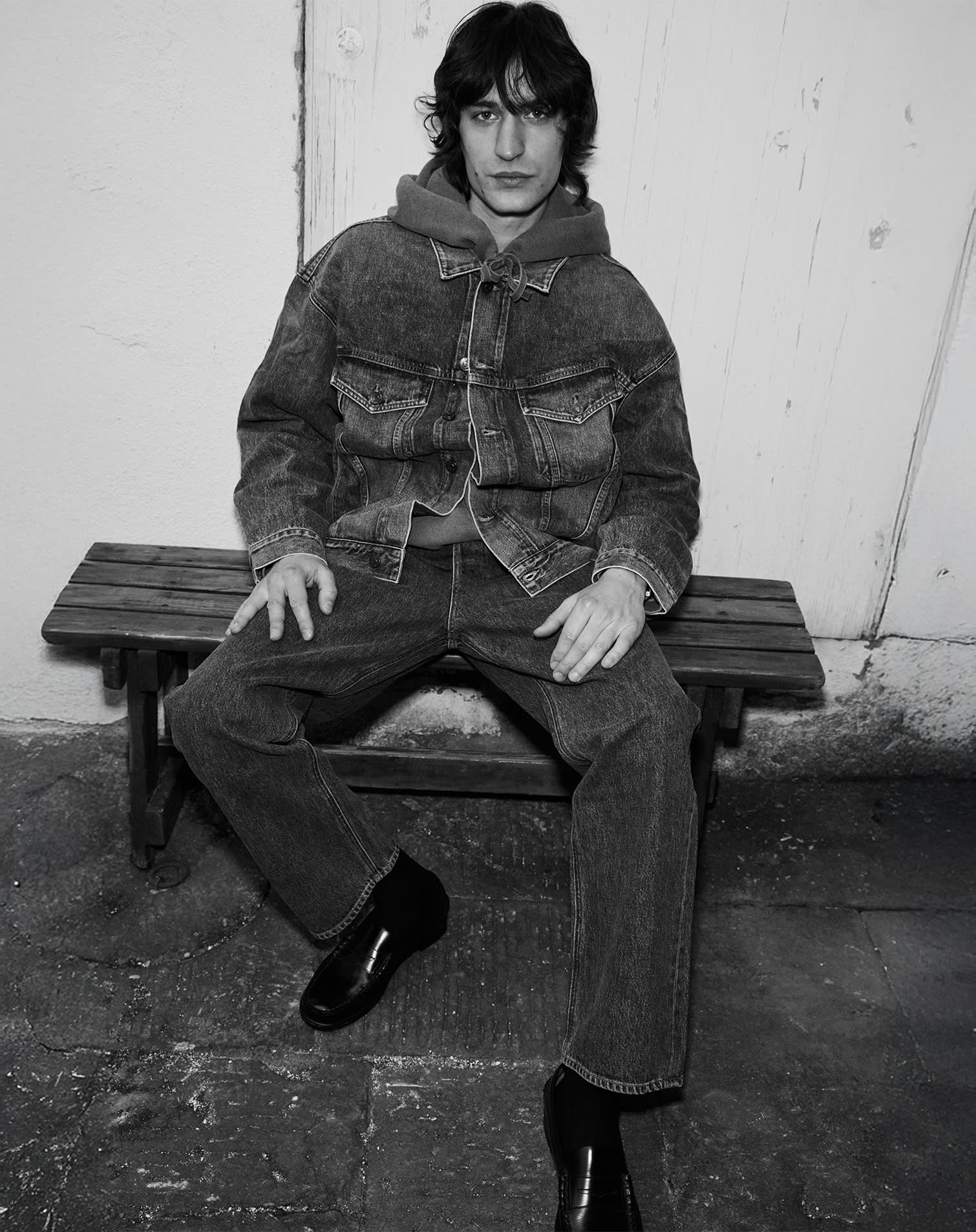 man wears jeans and denim jacket over hooded sweatshirt and black loafers