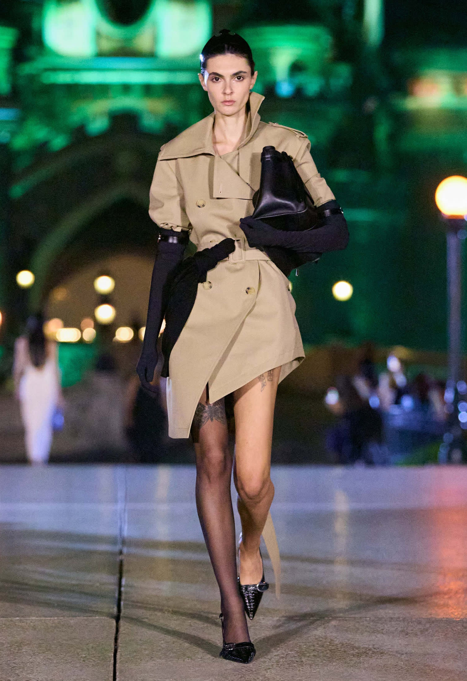 Weibliches Model auf dem Laufsteg in kurzem Trenchcoat mit schwarzer Tasche und schwarzen Pumps