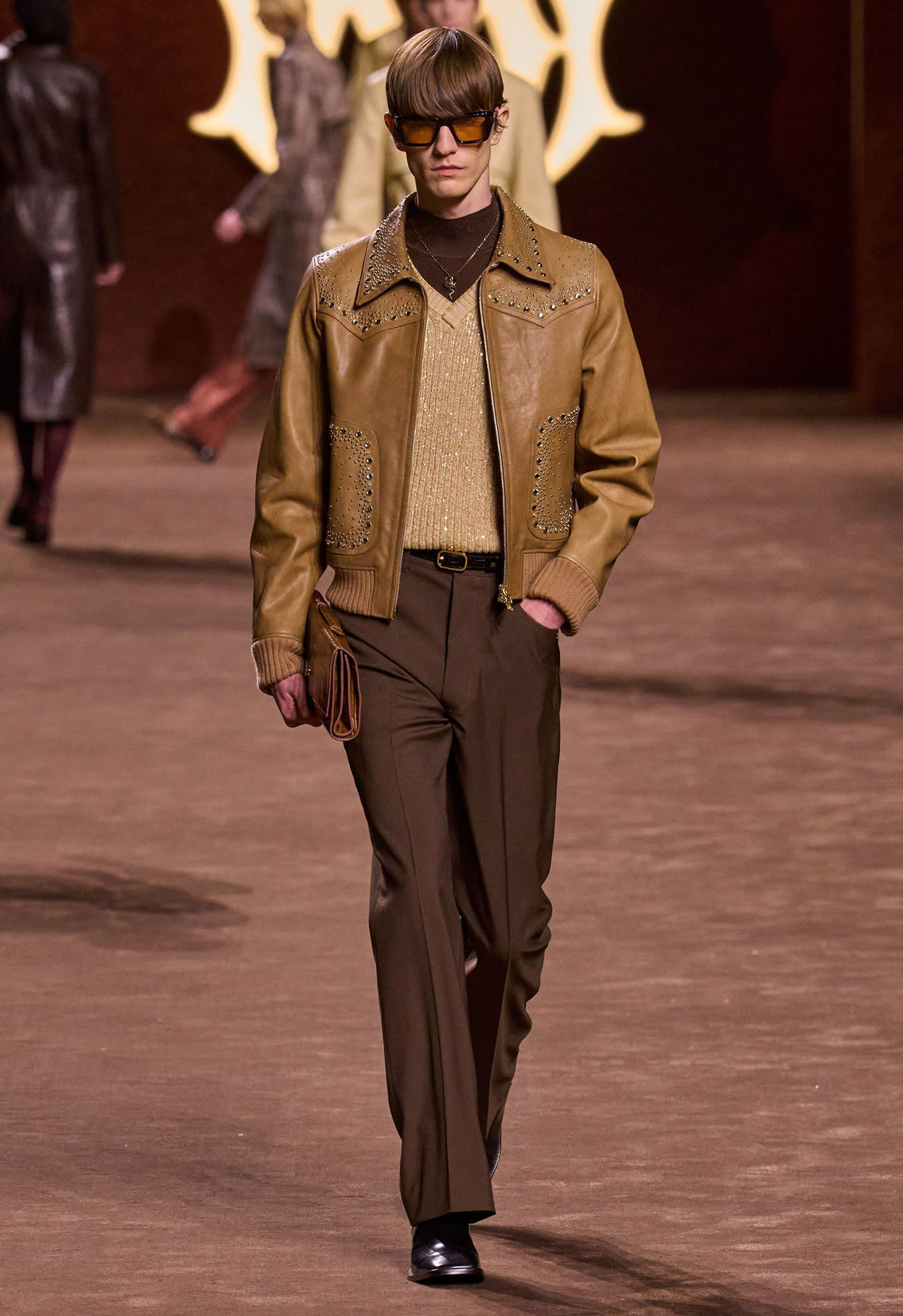 Studded brown leather jacket, gold knit sweater vest over dark brown turtleneck with brown pants and accessories