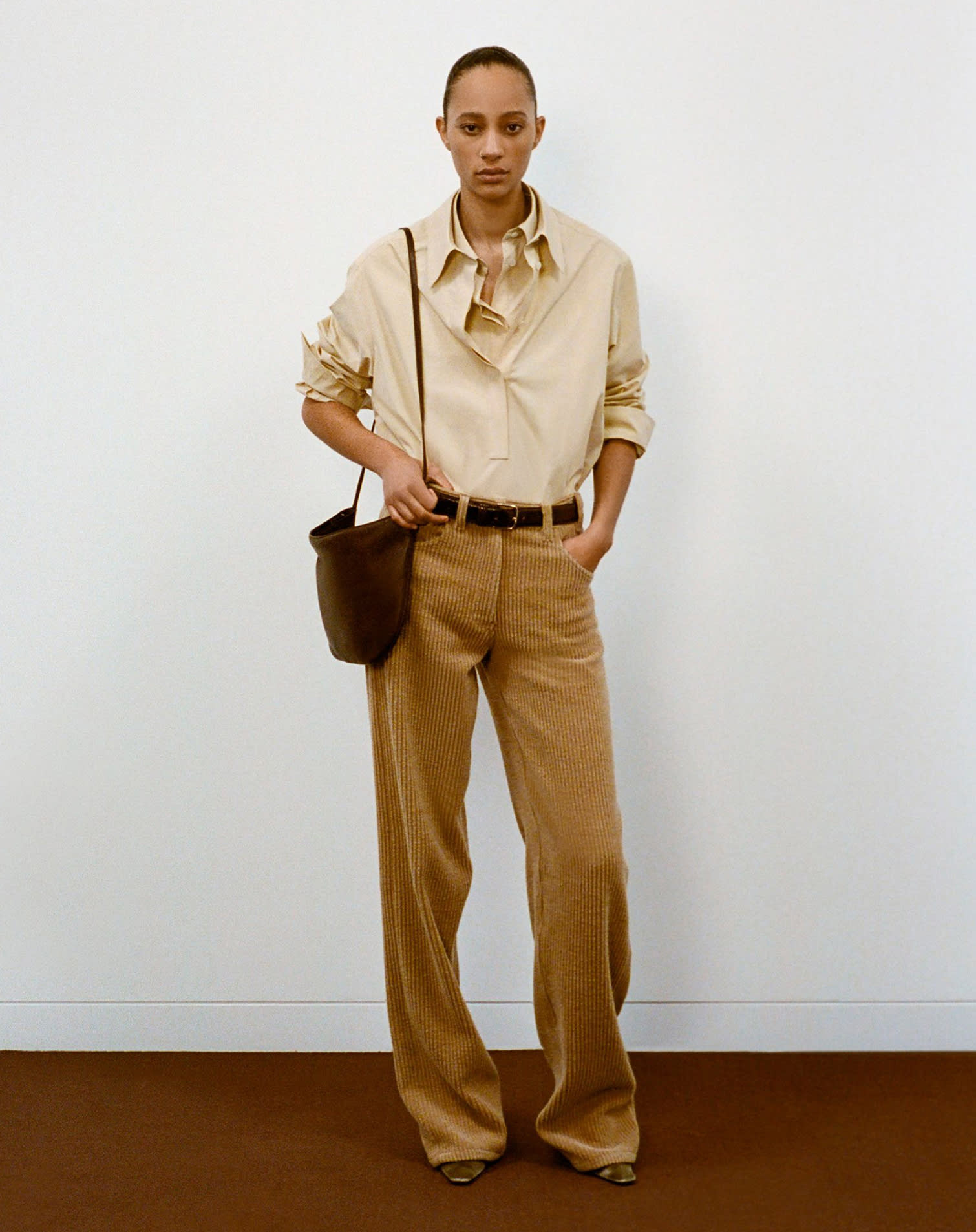 Female model wearing light brown corduroy pants, beige button-up shirt and dark brown leather bag and belt