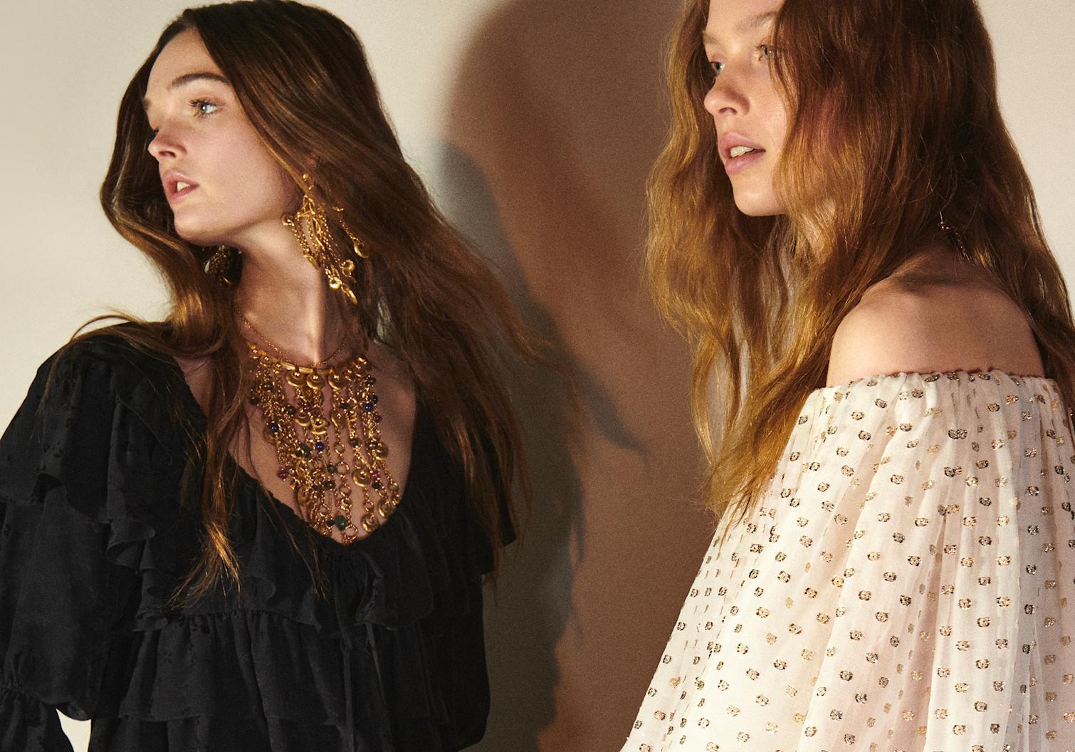 Two women wearing ruffled dresses—one in black with layered gold jewelry, the other in an off-shoulder ivory dress with gold embellishments—posed in warm, soft lighting